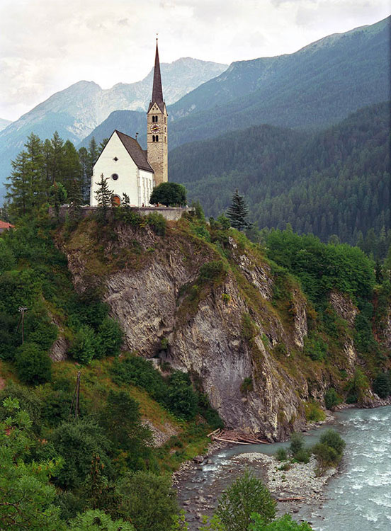 A church with a tall steeple sits on a bluff above a rocky stream in the Engadine 